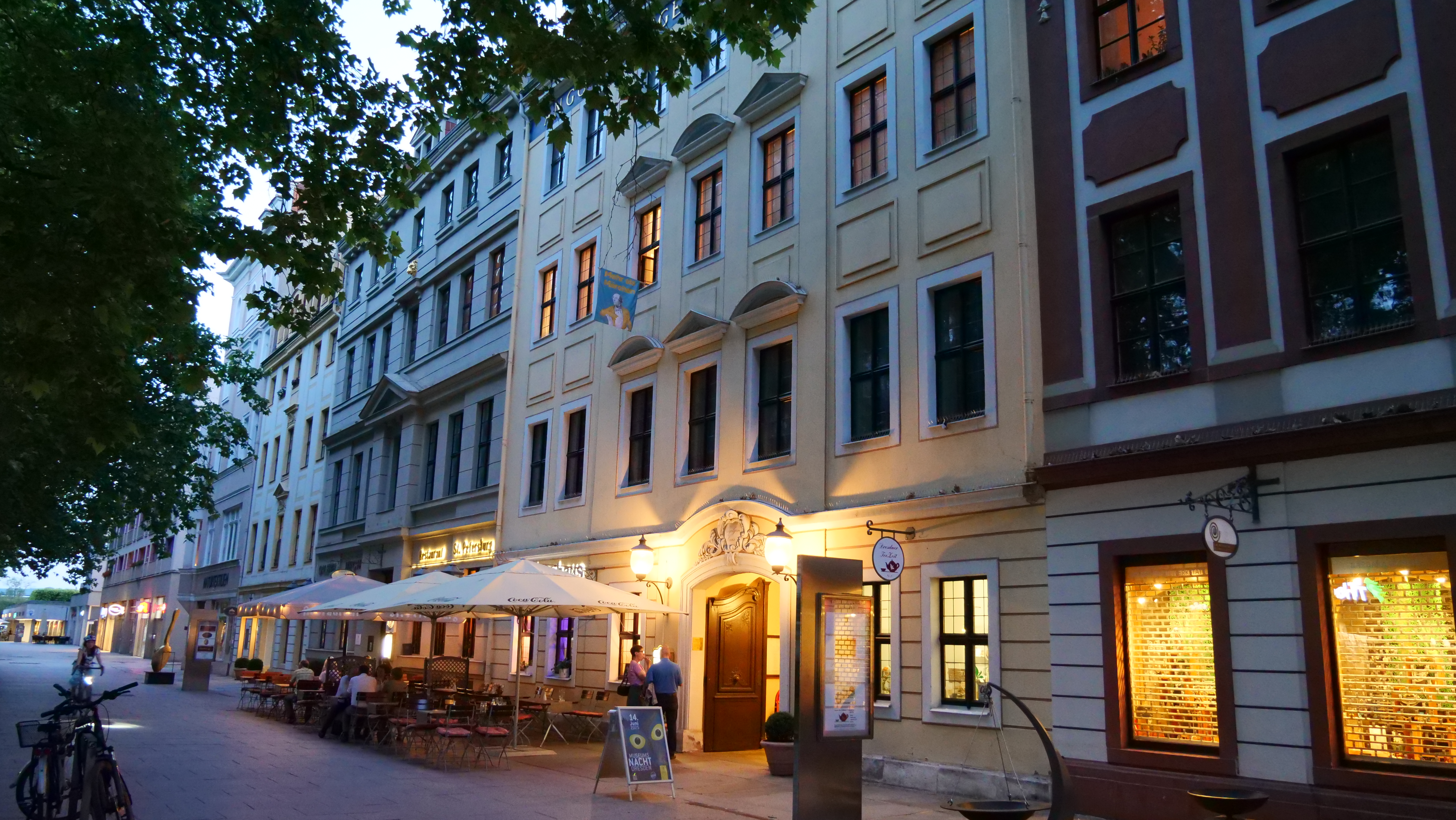 AI generated: The image shows a row of historic buildings with a restaurant or café on the ground floor, whose outdoor area is furnished with tables and parasols. The street is quiet and deserted, except for a few passers-by.