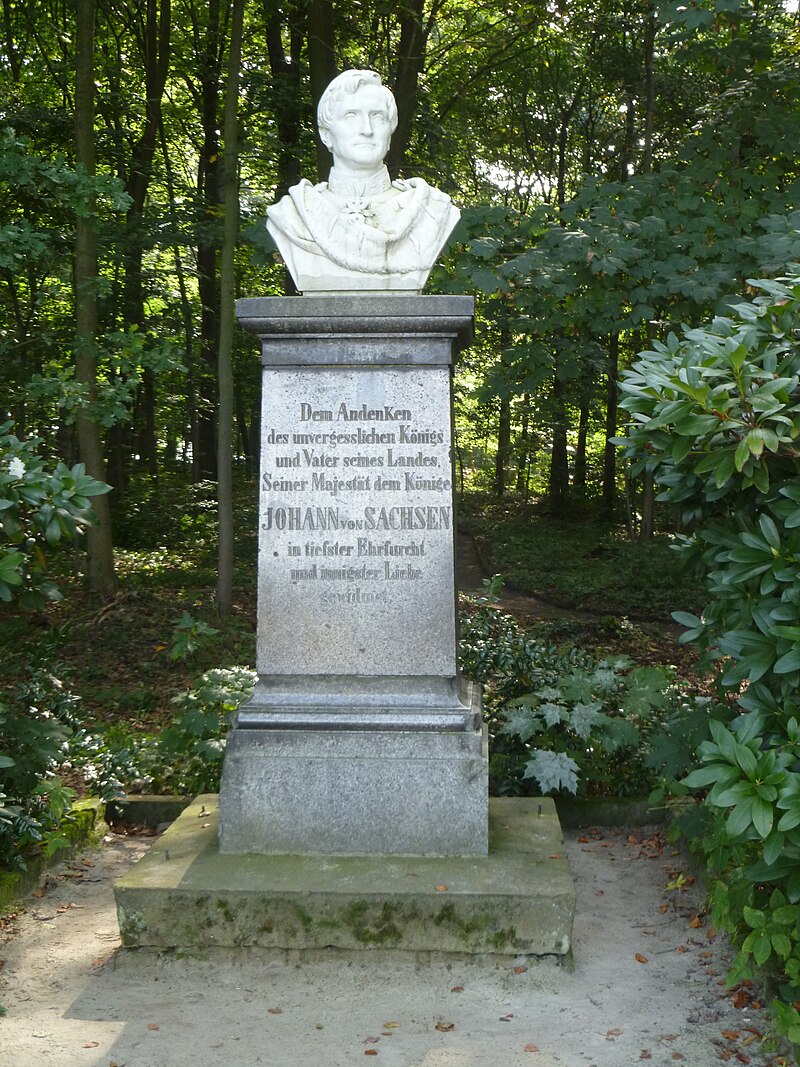 KI generiert: Das Bild zeigt eine Büste von Johann von Sachsen auf einem Sockel mit Inschrift, die an ihn erinnert. Die Statue steht in einem bewaldeten Park oder Garten.