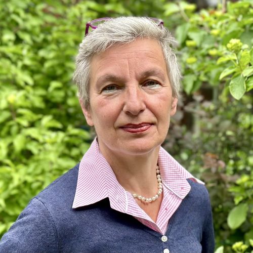 KI generiert: Das Bild zeigt eine Person mittleren Alters, die in einem Garten oder vor einer grünen Umgebung steht. Sie trägt eine Perlenkette und wirkt freundlich.