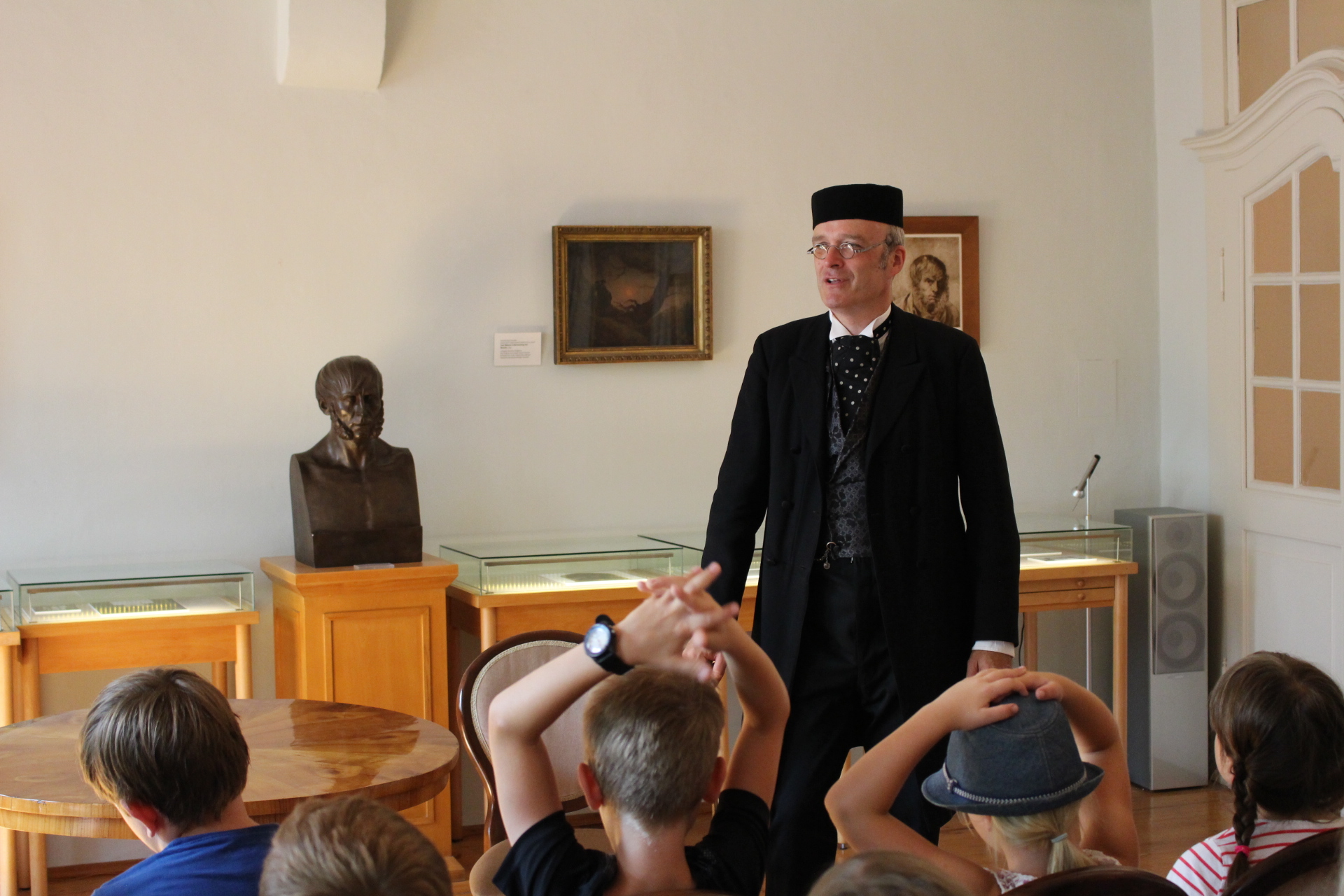 KI generiert: Ein Mann in historischer Kleidung spricht zu einer Gruppe von Kindern in einem Museum oder historischen Raum. Im Hintergrund sind Vitrinen und eine Büste zu sehen.