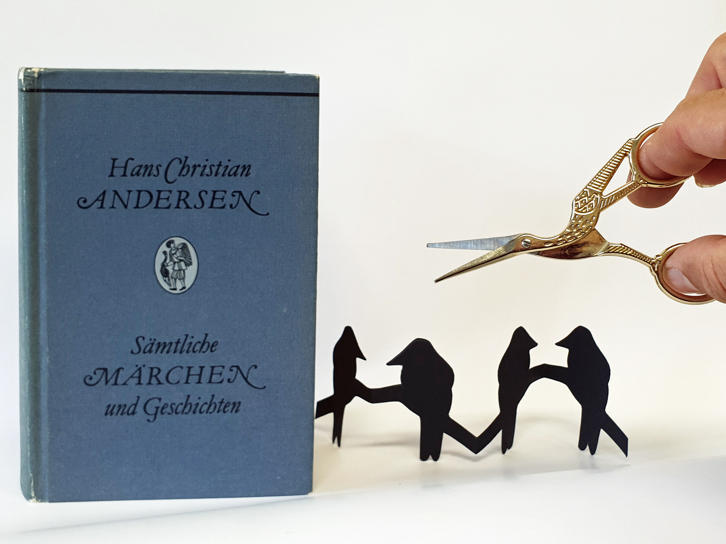 KI generiert: Ein Buch von Hans Christian Andersen steht neben einer Hand, die eine kleine Schere, geformt als Storch, hält. Dazwischen ist ein ausgeschnittenes schwarzes Papier zu sehen, das Figuren darstellt.