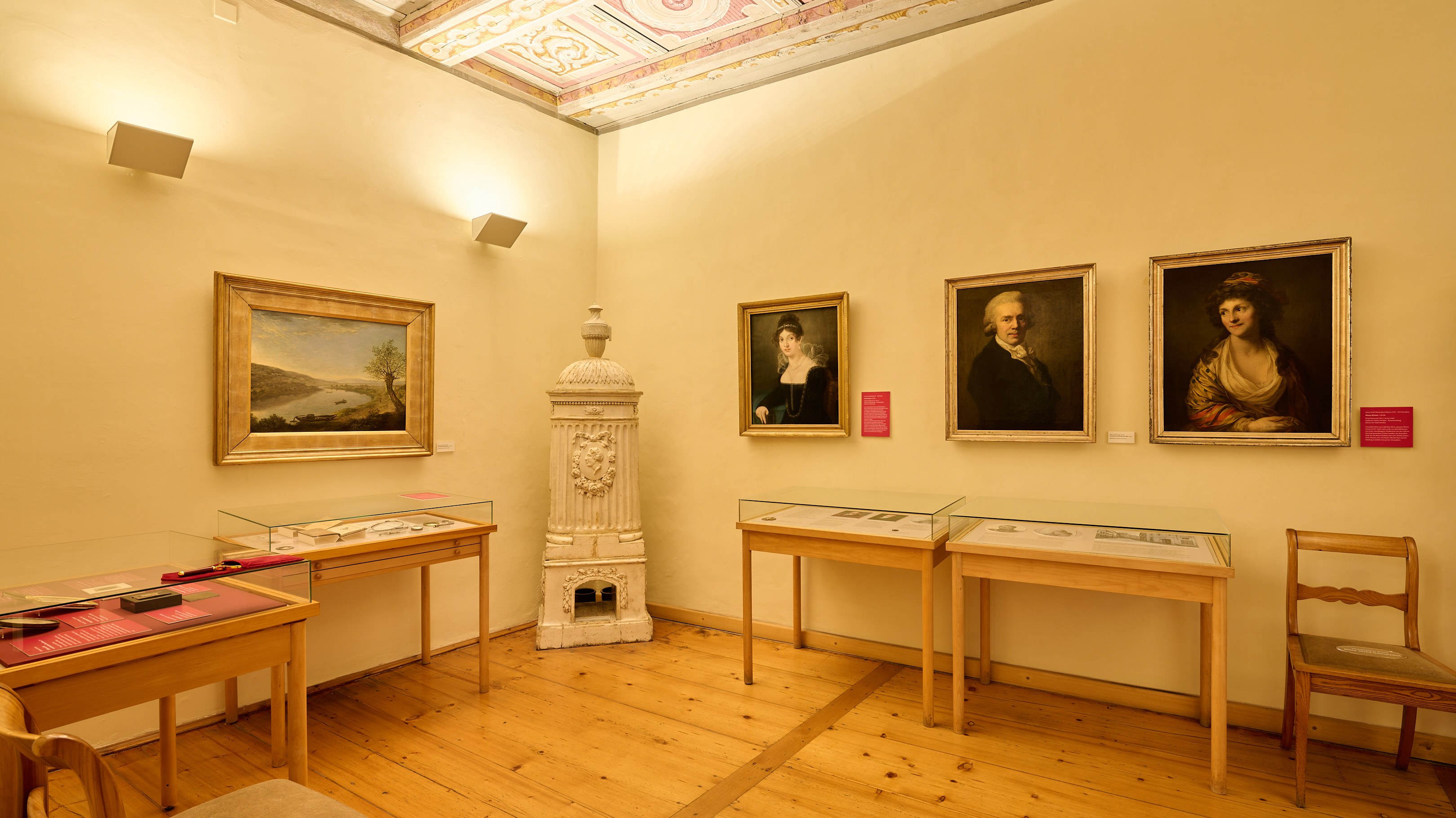 KI generiert: Das Bild zeigt einen Ausstellungsraum mit historischen Gemälden und Schautafeln, die in Glasvitrinen präsentiert werden. In der Mitte des Raumes steht ein dekorativer, antiker Ofen.